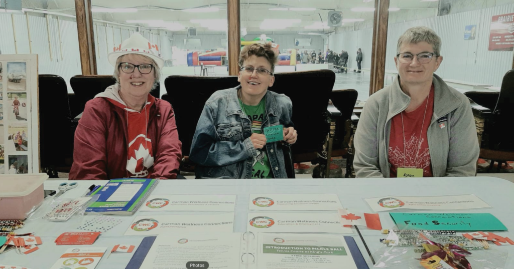 Three Carman Wellness Connections volunteers welcome visitors at a community event information table.
