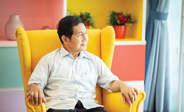 A man relaxes in a bright yellow armchair, looking toward a window in a colourful, comfortable living room.