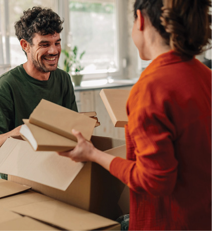 A smiling man and woman unpack cardboard boxes together in a bright home, sharing a moment while settling in.