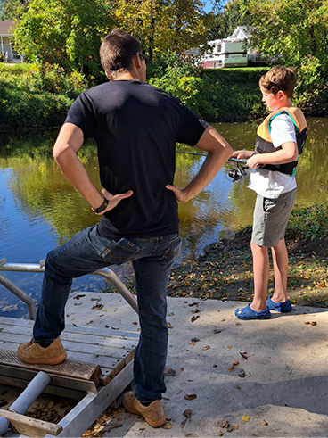 An adult and child stand beside a river, with the child holding a fishing rod during an outdoor activity.