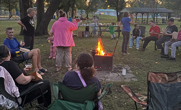 Community members gather around a bonfire in a park, sitting in lawn chairs, chatting and enjoying an outdoor evening together.