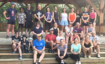 A group of adults and children pose together on wooden steps beneath a park shelter before a community recreation activity.