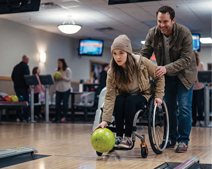 A woman in a wheelchair rolls a bowling ball down the lane while a man stands beside her offering encouragement.