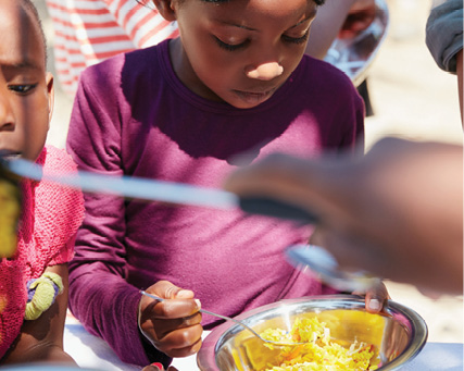 Children gather around a bowl of food, with one child holding a spoon during a shared meal.