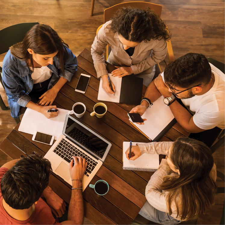 Five adults collaborate around a table with a laptop, notebooks and coffee, sharing ideas during a group meeting.