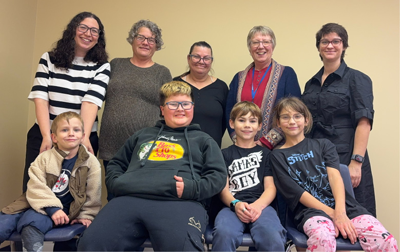 Five adults stand behind four children, smiling together for a group photo indoors.