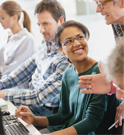 A smiling woman works at a computer while an instructor offers guidance during an adult learning session.