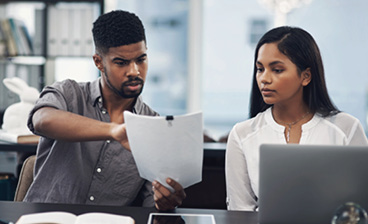 A man reviews paperwork with a woman seated beside him at a laptop, discussing information in an office setting.