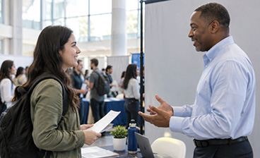 A woman holding documents speaks with an employer at a career fair, discussing employment opportunities.