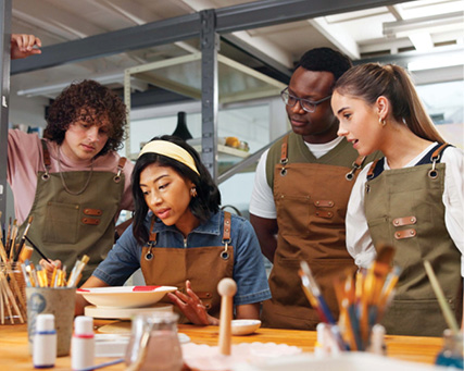 Four adults wearing aprons gather around a pottery project, learning and practising hands-on skills in a workshop.