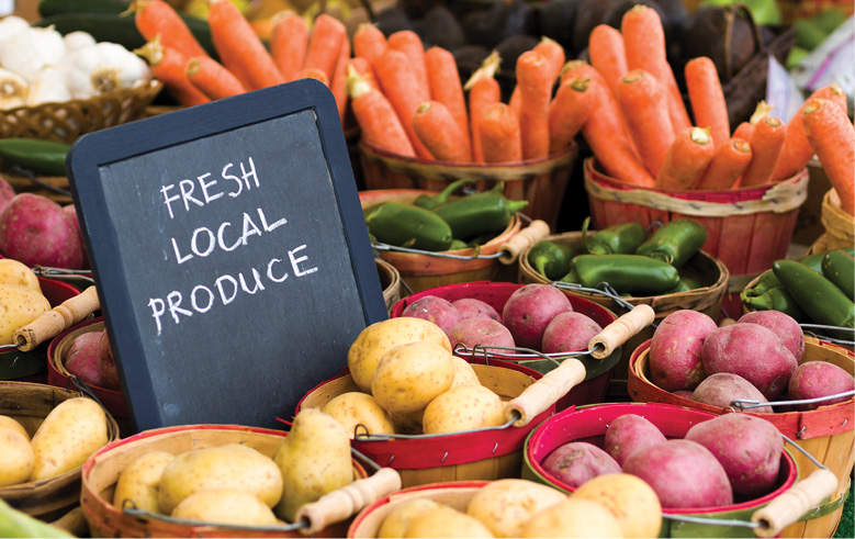 Baskets of fresh carrots, potatoes and peppers surround a chalkboard sign reading “Fresh Local Produce” at a market.