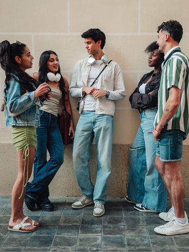 Five young adults stand together, smiling and chatting in a relaxed social setting.