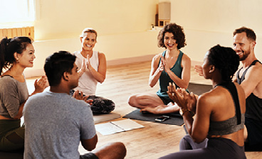 Six adults sit in a circle on the floor, smiling and clapping during a group wellness activity.